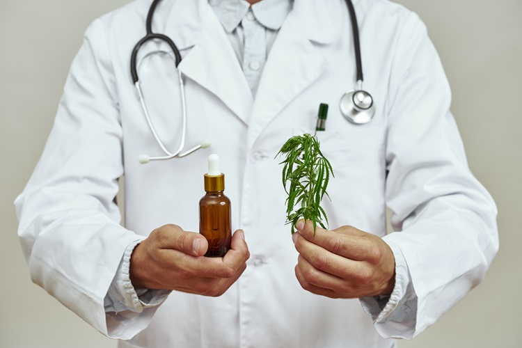 Imagem de um homem de bata branca segurando uma folha de marijuana e um frasco de óleo essencial de canabinóide