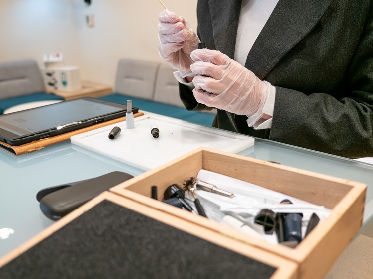 Une image en gros plan d'une personne portant des gants assemblant ou travaillant avec des composants de vapotage sur une table en verre. La personne manipule de petites pièces, peut-être une cartouche de vapotage ou un embout buccal, à l'aide d'un outil. Au premier plan, il y a une boîte en bois ouverte contenant divers outils et accessoires liés à la vape. Une tablette et d'autres appareils électroniques sont également visibles sur la table, indiquant un espace de travail axé sur l'assemblage ou la réparation de la vape.